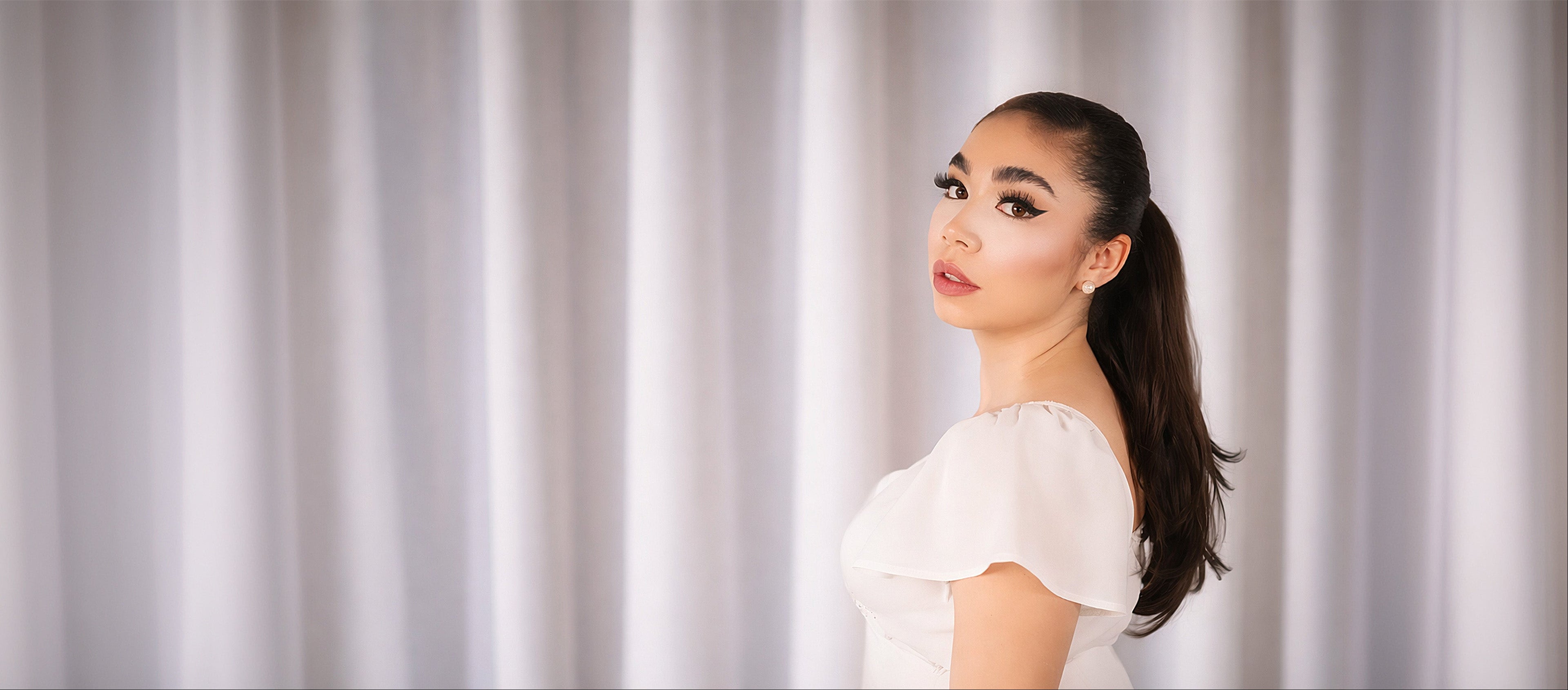 Woman in a white blouse standing against a plain curtain background